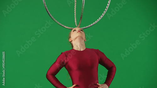 Foto Young circus gymnast in a red leotard standing on a green background and holding the hoop on her forehead