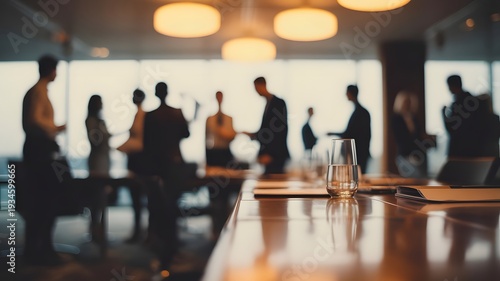 A group of business professionals networking and socializing in a modern conference room with a sleek table and glass of water in the foreground.