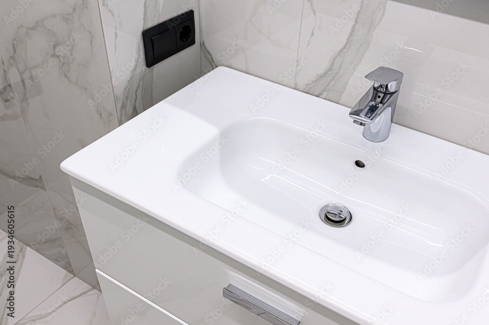Fototapeta premium A sleek white ceramic sink with a chrome faucet is mounted on a white vanity cabinet; set against marble-patterned tiles with a black wall outlet nearby.