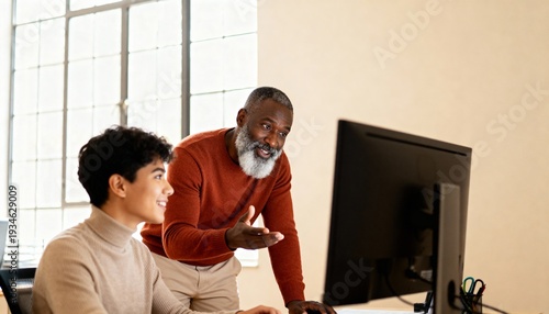 Senior Black man mentoring a young man at a computer in a bright office. Professional collaboration and teaching concept. Older male mentor guiding a student in a workplace setting