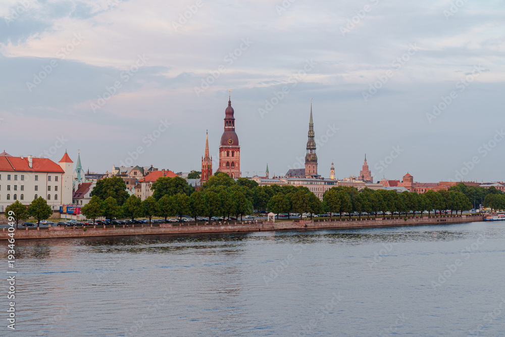 Obraz premium Warm sunset bathes historic buildings in Riga Latvia beside calm river Duna. Trees line the waterfront, framing the skyline softly. Red-tiled roofs and spires glow in golden light Panorama