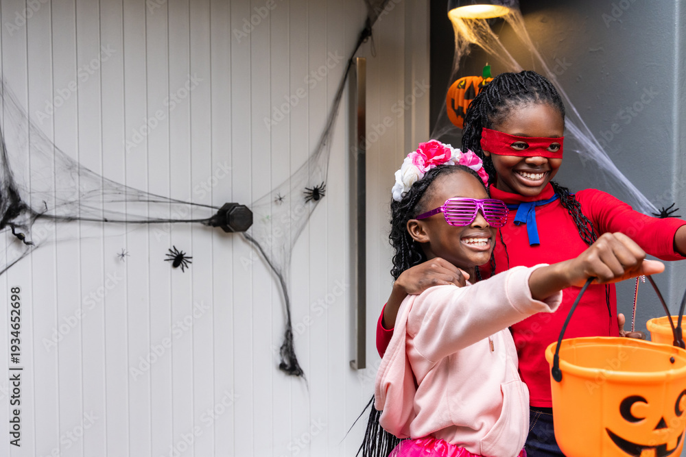 Naklejka premium Two African American girls posing at front door amid faux webs, holding pumpkin buckets, copy space
