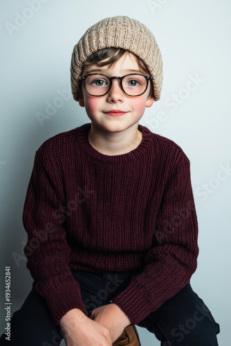 Young boy wearing glasses and a knit hat poses in a cozy sweater against a light background