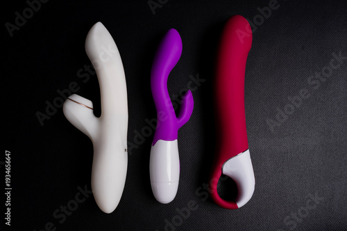 Fotografie White suction vibrator with clitoral stimulator, purple rabbit vibrator and red phallic vibrator placed on dark textured background under studio lighting