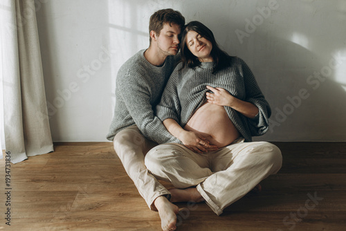 Stylish pregnant couple hugging baby bump and sitting on floor. Fertility, healthy pregnancy and expecting new life concept. Happy Family in cozy sweaters and jeans holding pregnant belly