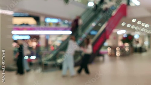 Blurred bokeh background footage of a modern shopping mall interior, with anonymous visitors walking through common areas and moving on an automated escalator connecting different floors.