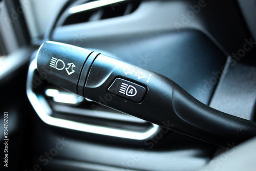 Close-up of the black multifunctional turn signal lever on the steering column. Glossy plastic lever with white icons for headlights, high beams, turn signals, and fog lights, built-in lane change ass