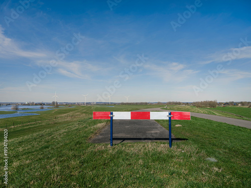 Tableau sur toile Red and white road barrier blocking dike road with flooded polder and wind turbi