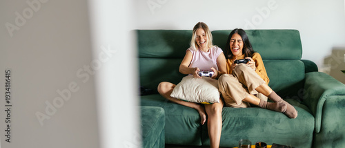Schilderij op canvas Women friends sharing a fun moment gaming at home, relaxing on a green sofa