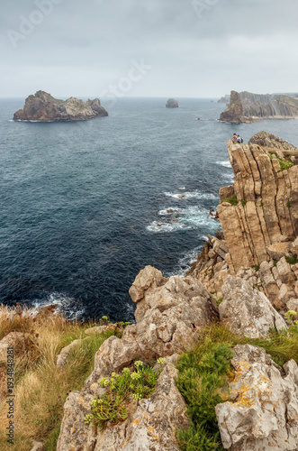 Sheer cliff over the sea on the Costa Quebrada, Cantabria, Spain, with rock formations in the sea — the Urros de Liencres — in the background.
