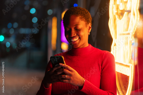 Smiling young black woman using smartphone beside glowing neon lights on city street at night. Happy female in red sweater text messaging on mobile phone