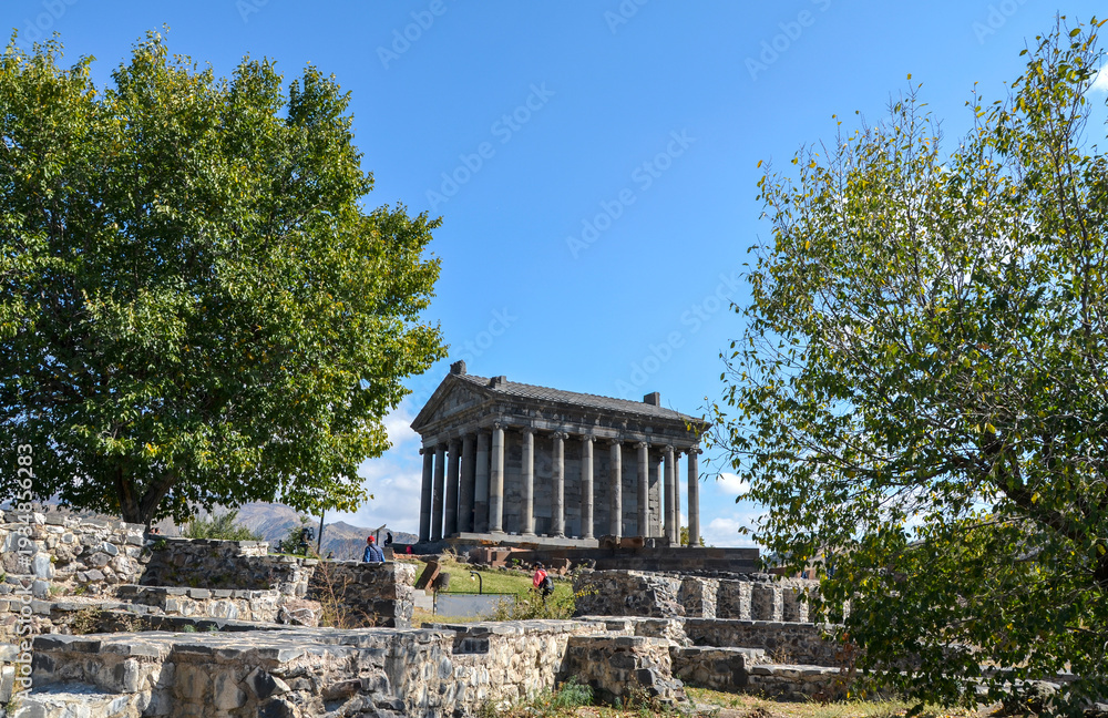 Fototapeta premium Ancient stone Garni Temple with classic columns stands on a grassy hill under a blue sky. Rugged mountains rise in the distant background. Armenia