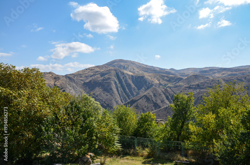 Rolling hills under a blue sky with fluffy clouds. Green trees frame the foreground, overlooking a vast, arid valley. Sunlight illuminates the dry, textured mountain slopes. Armenia