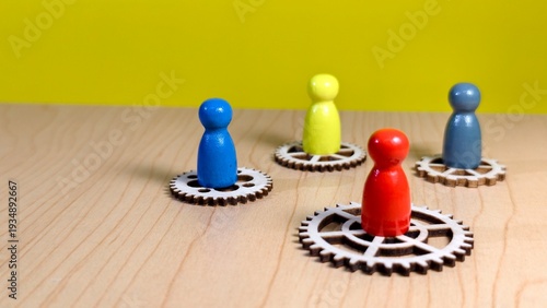 Photos Four colorful wooden game pieces stand atop individual gear wheels on a wooden surface, symbolizing teamwork, mechanical synergy, and organizational roles against a solid yellow backdrop