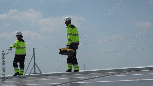 Solar energy specialists in high-visibility gear walking on rooftop during site inspection, Maintenance team with toolbox performing routine check on photovoltaic system