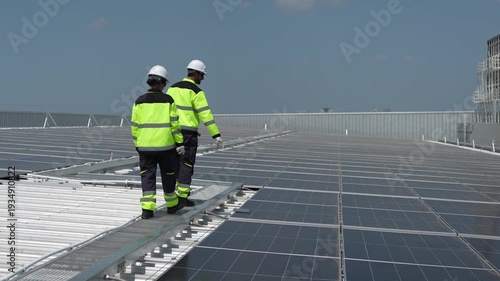 Engineers in high-visibility uniforms walking on rooftop walkway of large solar farm, Technical team pointing at solar panels during routine inspection on industrial roof