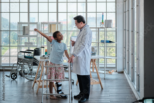 Professional senior Asian doctor training walker, helping disabled Black girl to walk, leg fracture recovery, tenderness encouraged with smile in orthopedic ward, and pediatric healthcare hospital.