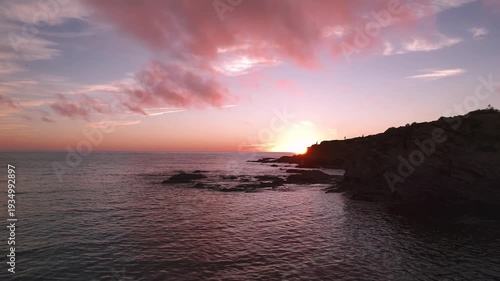 Atlantic sun sets behind rugged coastal cliffs near Porto Covo along Costa Vicentina in Alentejo Portugal casting warm light across ocean surface, drone glide.
