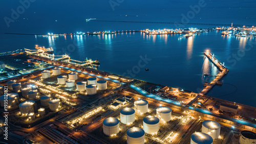 Fotografie Industrial oil terminal harbor at twilight with illuminated refinery tanks, cargo docks, and shipping infrastructure reflecting on calm sea, capturing global energy logistics and maritime industry
