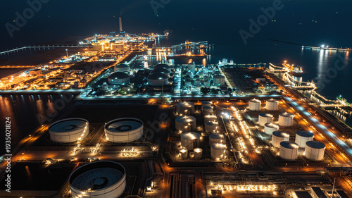 Fotografie Night aerial panorama of coastal petroleum refinery complex with glowing storage tanks, pipelines, and shipping port infrastructure reflecting on dark sea, representing modern energy industry