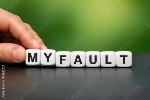 Admitting Responsibility: A hand carefully aligns word dice spelling out MY FAULT, against a blurred green backdrop, highlighting the concept of personal accountability.