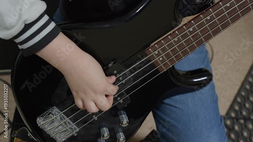 Close-up video of a young child's hand playing a black electric bass guitar. The kid plucks the strings, practicing music. Focus on musical education and learning an instrument.