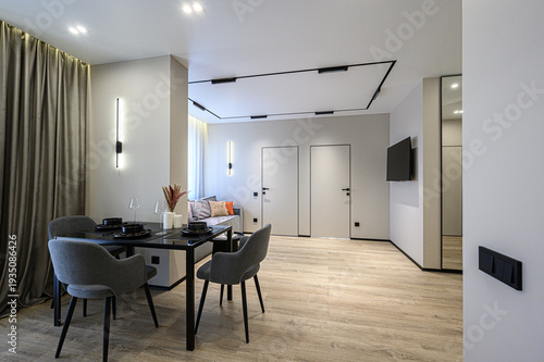 A modern minimalist living space with a black dining table, grey upholstered chairs, neutral curtains, hidden doors, track lighting, light wood flooring, and a mounted TV.