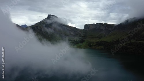 Dramatic mountain landscape with low clouds and a serene lake at Barrage de Roselend in the French Alps. Moody atmosphere with rugged peaks and lush slopes.