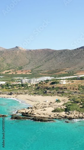 Aerial view of Falasarna beach with turquoise water, rocky coastline, and mountains in Crete, Greece. Clear blue sky and natural Mediterranean landscape.