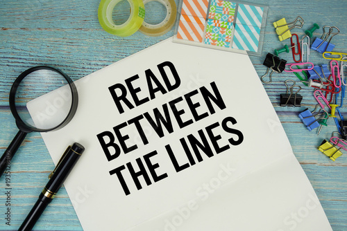 фотографія Read Between The Lines text on a paper surrounded by office supplies and magnify