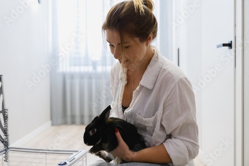 Close view of young woman holding her pet rabbit near an open cage in a bright modern apartment. Caring for a domestic animal in a calm home environment.