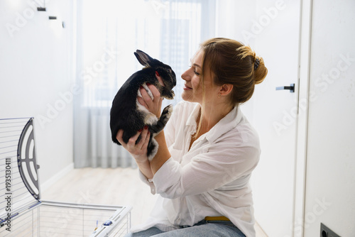 Happy young woman holding and cuddling her pet rabbit near an open cage in a bright modern apartment. Warm moment of affection between owner and domestic animal.