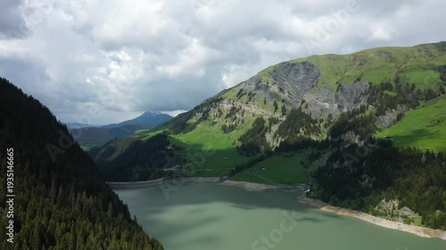 Scenic aerial view of Barrage de Saint Guerin dam surrounded by lush green mountains and forest under a cloudy sky in the French Alps.