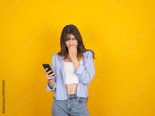 Young woman looking worried while checking smartphone, biting nails in anxiety. Female using mobile phone isolated on yellow background with copy space, concept of stress, bad news, social media or no