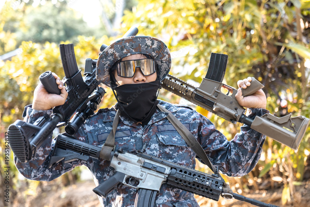 Obraz premium Confident soldier in full camouflage uniform and mask holding multiple assault rifles. An armed man with gun and tactical gear poses outdoors in forest for combat mission