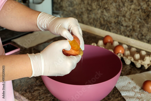 Gloved hands cracking a fresh brown egg into a pink mixing bowl on a kitchen countertop during food preparation, hygienic cooking process for baking homemade pastry or breakfast dishes