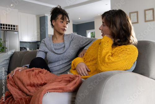 Female friends sitting on gray sofa at home, wearing yellow and gray sweaters with orange throw