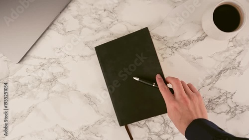 A hand holds a pen over a black notebook on a white marble desk. A silver laptop and a warm cup of coffee are also present, creating an organized and productive workspace