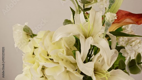 Observe an exquisite grouping of fresh white lilies, fluffy cream hydrangeas, and hints of green foliage. A single reddish-orange calla adds a touch of warm color