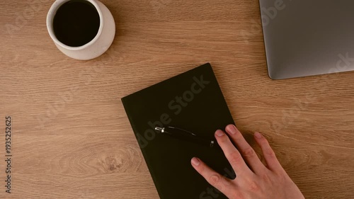 A person's hand extends over a natural wood desk, positioned near a white coffee cup, a black notebook, and a silver laptop. This shows a tidy workspace setup for productivity