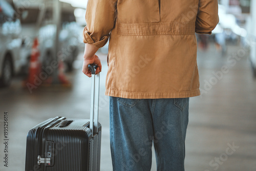 Back view of a young traveler pulling a black suitcase while walking through an airport or transport terminal during the day, representing modern mobility, global travel trends, journey