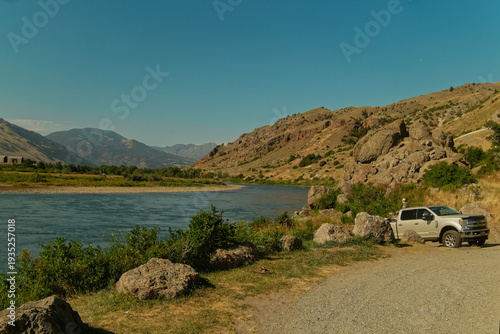 Vehicle By River. Lone Road Traveler Unloads Gear Beside Mountain Stream Under Bright Midday Sun