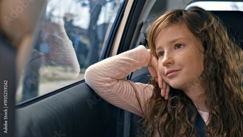 Inside a car, a young girl with long curly hair rests her head on her hand, gazing thoughtfully out the window. She appears relaxed, suggesting a casual, introspective moment during a road trip.