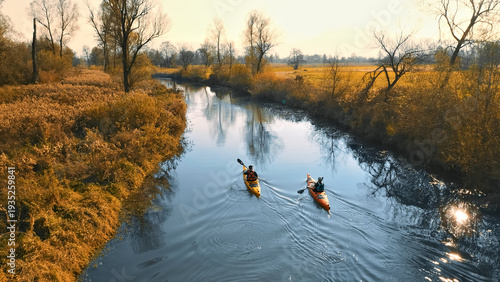Two kayakers navigate a serene, sunlit river surrounded by autumn foliage. The calm water reflects golden hues, creating a tranquil scene.