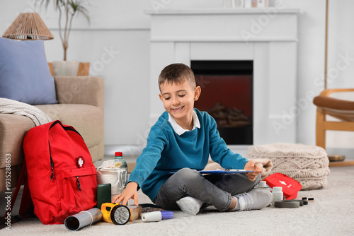 Fotografia Little boy with check-list packing emergency backpack on floor at home