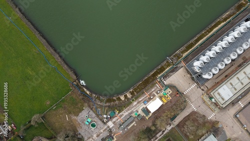 Aerial view of a large body of water, contrasted by the adjacent industrial structures and green fields, casting long shadows under the diffused light, Birmingham, England, United Kingdom.