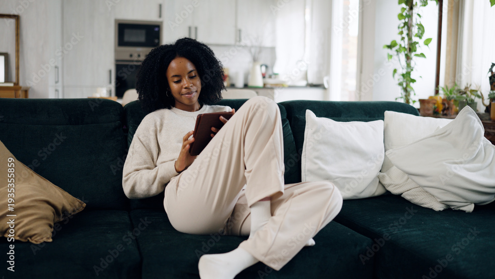 Obraz premium Young black woman relaxing on a comfortable sofa at home, casually using a digital tablet for entertainment and connectivity