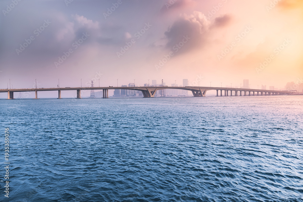Obraz premium bridge providing a modern architectural link over calm water during an atmospheric morning in Abu Dhabi. Urban silhouettes visible in the distance