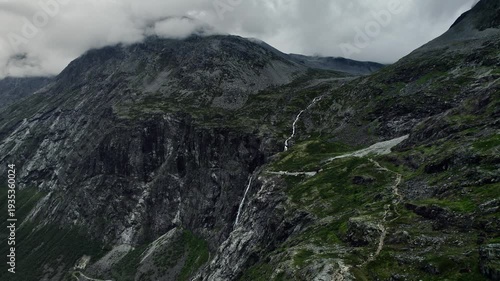 Drone descending near steep cliffs showcasing Trollfossen waterfall and Storgrovfjellet mountain. Clouds cover the peaks as the path leads through the rugged terrain in the natural landscape of Norway