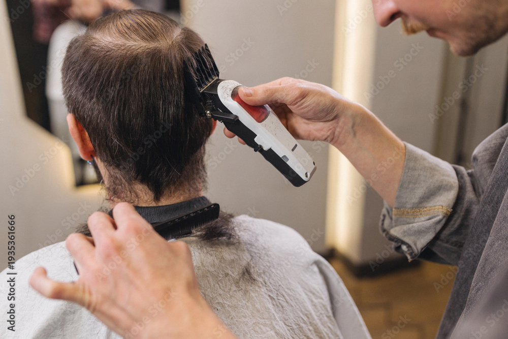 Fototapeta premium Professional barber trimming client hair with electric clipper in modern barbershop. Male grooming service, precise hairstyling process, focused expression, contemporary salon interior.